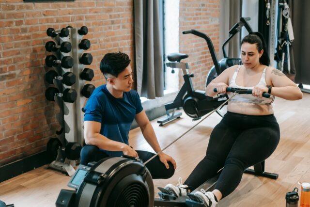 Image of a personal trainer providing guidance and support to a larger woman as she uses the machine rower during a workout session, fostering inclusivity and encouragement in fitness