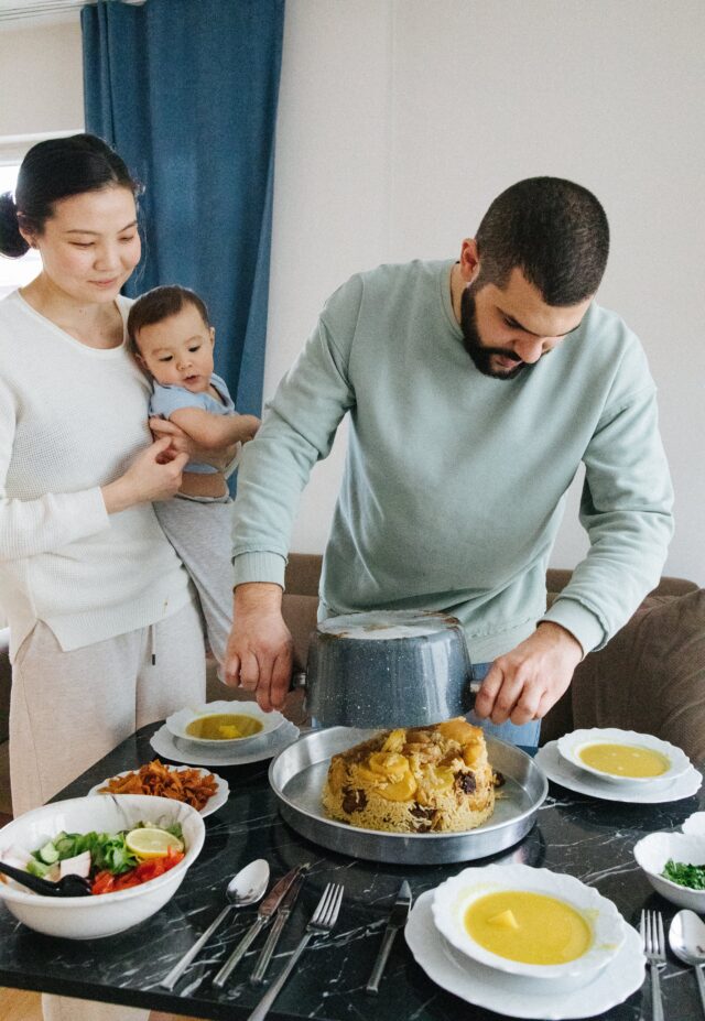 Happy wife holding child while man prepares food, capturing a heartwarming moment of family joy and domestic teamwork.