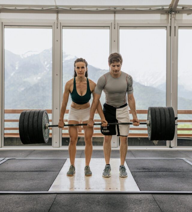 Man and woman combining their efforts to lift a barbell loaded with weights, exerting full strength and teamwork in the challenging lift.