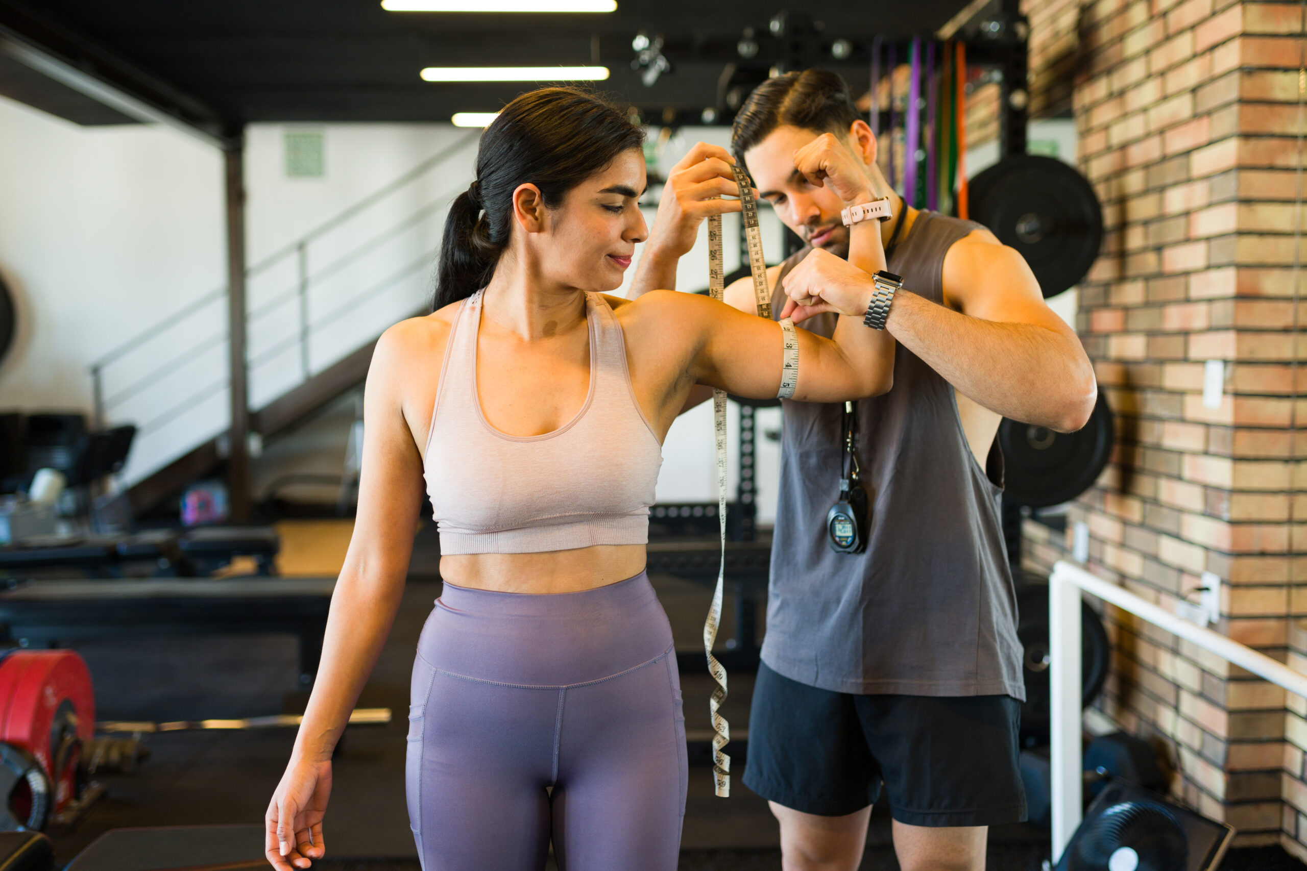 Professional trainer measuring female client's biceps with tape measure during fitness evaluation at health club