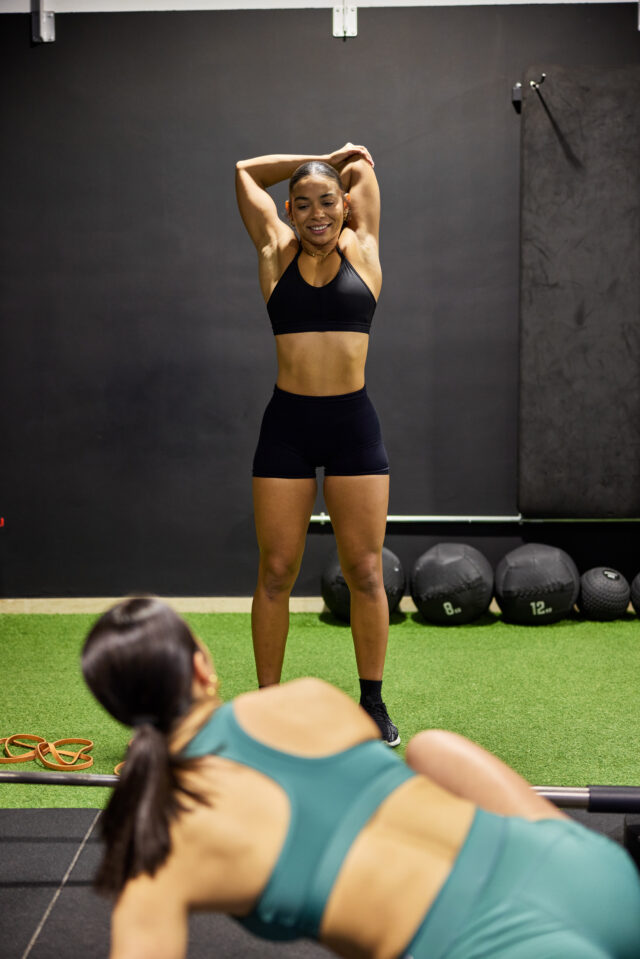Two women are engaged in a workout at a gym. One woman is performing an exercise while the other stretches. Gym equipment is visible in the background.